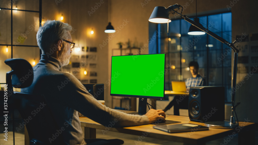 Aufkleber Creative Middle Aged Designer Sitting at His Desk Uses Desktop Computer with Green Mock-up Screen. In the Evening Two Creative Employees Working on Computers in the Office with Cityscape Window View
