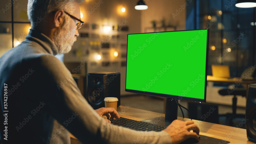 Poster Creative Middle Aged Designer Sitting at His Desk Uses Desktop Computer with Green Mock-up Screen. In the Evening Creative Employe Working on Computers in the Office with Cityscape Window View