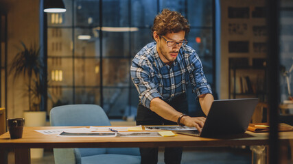 Young Creative Designer Working at His Desk, Focused on the Laptop and Dockuments, Choosing Right Design, Concept and Sketches. In the Background Evening Office Studio with Cityscape Window View