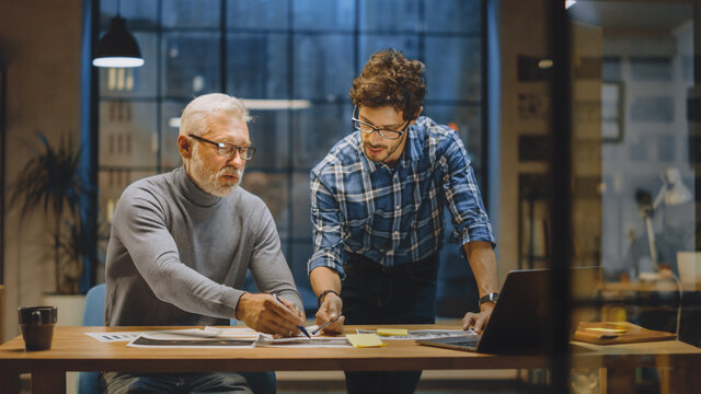 Chief Creative Designer Discusses Design Sheets with Young Developer. Two Professionals Having Discussion, Working with Documents, Choosing Right Concept. Evening in the Stylish Modern Studio Office