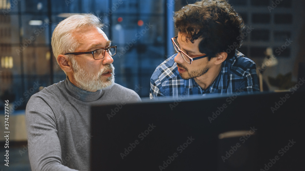 Poster Chief Creative Designer Shows Desktop Computer Screen to a Young Developer, They Discuss Project. Two Professionals Having Discussion. Choosing Right Design Concept. Evening in the Studio Office