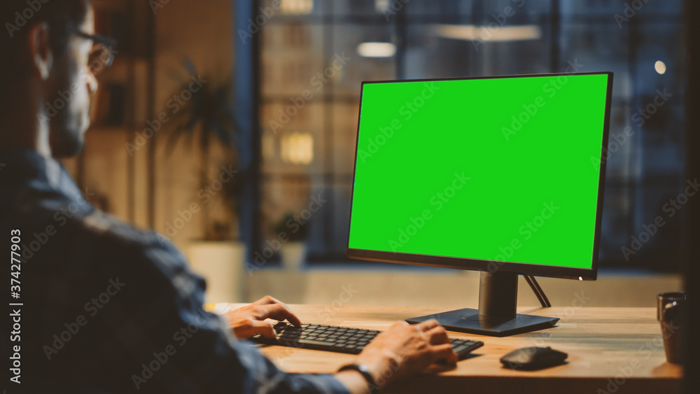 Poster Over the Shoulder: Creative Young Man Sitting at His Desk Using Desktop Computer with Mock-up Green Screen. Evening in the Stylish Office Studio with City Window View