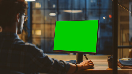 Over the Shoulder: Creative Young Man Sitting at His Desk Using Desktop Computer with Mock-up Green Screen. Evening in the Stylish Office Studio with City Window View