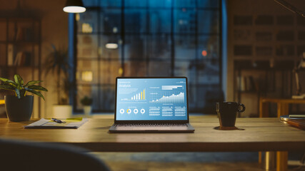 Shot of a Laptop on a Desk with Statistics Graph, Chart and Various Data, Showing Company Growth and Success. In the Background Warm Evening Lighting and Open Space Studio with City Window View.