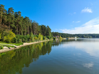 Landscape of the river Bank with bushes, trees and a small beach. Forest along the river with a reflection in the water against the bright blue sky.