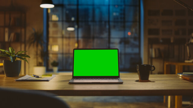 Mock-up Green Screen Laptop Standing on the Desk in the Modern Creative Office. In the Background Warm Evening Lighting and Open Space Studio with City Window View.