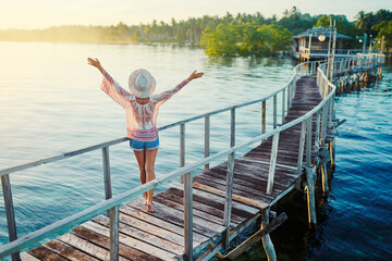 Fototapeta na wymiar Feel freedom! Vacation on tropical island. Back view of young woman in hat rising hands enjoying sunset sea view from wooden bridge terrace, Siargao Philippines.
