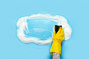 Hand in a yellow rubber glove holds a cleaning sponge and wipes a soapy foam on a blue background. Cleaning concept, cleaning service. Banner. Flat lay, top view - obrazy, fototapety, plakaty
