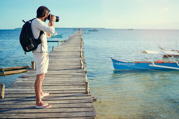 Photography and travel. Young man with rucksack standing on wooden fishing pier taking photo of beautiful tropical sea view.