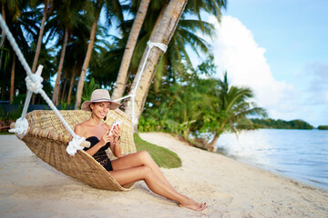 Vacation and technology. Young pretty woman in hat and swimsuit using smartphone laying in hammock on the beach.