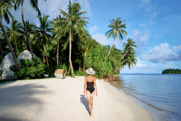 Vacation on the seashore. Back view of young woman walking away on the beautiful tropical white sand beach.