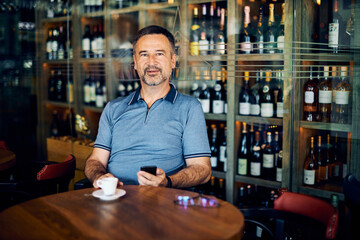 Senior adult sitting in winery, using smart phone on a coffee break while looking at camera.