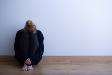 Sad teenager girl sitting curled up on the floor, copy space on empty wall