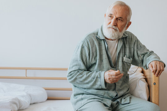 elderly man with gray beard and hair sits in pajamas on the bed with glasses in hand - Powered by Adobe
