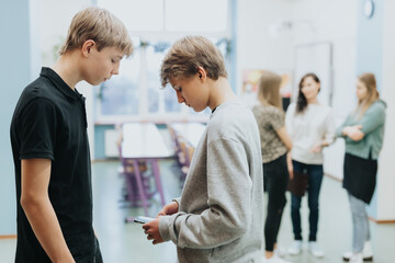 Obraz premium Two teenagers are standing together at the side of the classroom, one of them is checking something on a mobile phone