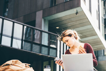 Сoncentrated blonde girl student types on modern laptop eating tasty sandwich on bench in city.