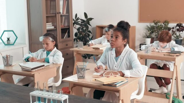 Handheld tracking shot of cute girls and boys in lab coats sitting at desks in classroom and writing in their notebooks while learning chemistry
