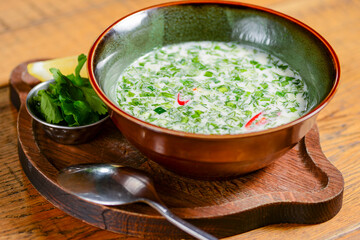 Popular spring and summer cold soup okroshka. Okroshka in a bowl, lemon and herbs on rustic wooden table.