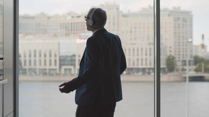 Cheerful mature businessman standing near large window listening to music in headphones and dancing