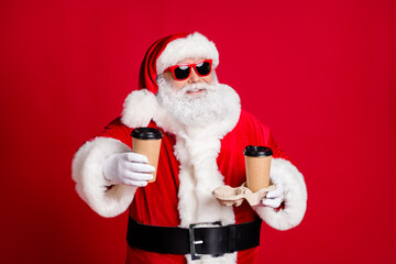 Portrait of nice handsome cheerful cheery bearded Santa holding in hand paper cups latte inviting giving you share friend delivery service isolated bright vivid shine vibrant red color background