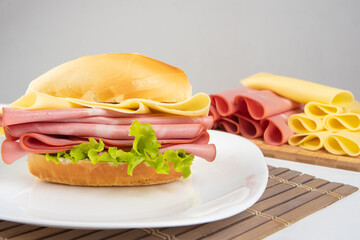 Mortadella sandwich, lettuce and cheese on a white plate on a mat and slices of mortadella and cheese next to it on a cold board on a table with white towel, selective focus. © Milton Buzon