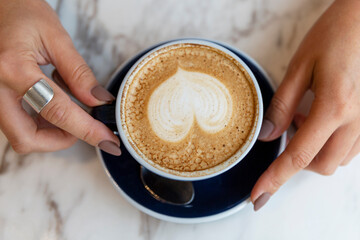 Cappuccino with heart-shaped foam in a blue cup on a marble table surface in female hands. Top view.