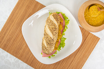 Mortadella and lettuce sandwich with oregano-covered bread, on a white plate and a bamboo mat and pepper sauce and mustard on a table with white towel, top view.