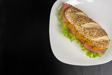 Mortadella and lettuce sandwich with oregano-covered bread, on a white plate and pepper sauce and mustard on a table with black towel, Selective focus.