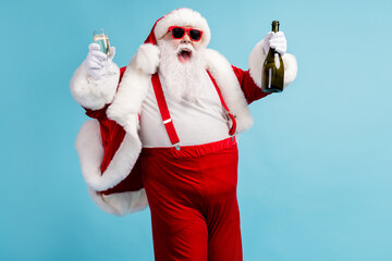 Fototapeta na wymiar Photo of retired old man grey beard funky hold bottle full glass sparkling wine can't stop drink crazy open mouth wear santa x-mas costume suspender sunglass cap isolated blue color background
