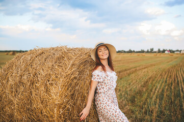 Naklejka na ściany i meble Woman in a summer dress and straw hat poses on a haystack. Wheat field and blue sky at sunset.