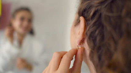 Close up of smiling young woman putting on earring and looking to mirror at home bathroom