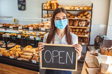 Portrait of a happy business owner with an open sign on the bakery shop and smiling - food and drinks concepts. Woman working at a bakery wearing a facemask to avoid the coronavirus