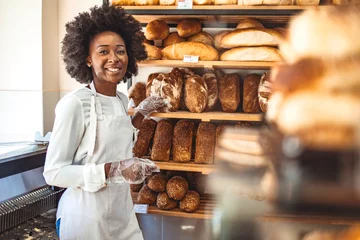 Papier Peint photo Lavable Boulangerie Smiling baker woman standing with fresh bread at bakery. Happy african woman standing in her bake shop and looking at camera.Baker with breads in background. Beautiful and smiling woman at bakery shop