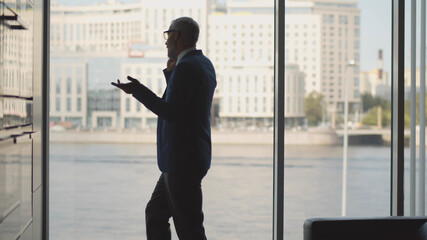 Concentrated businessman wearing suit talking on mobile phone in modern office near large window