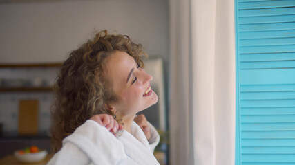 Pretty woman wearing bathrobe and standing near window while yawning after waking up.