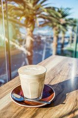 A glass of coffee latte on a wooden table with tropical nature background - palms and the ocean on a sunny day