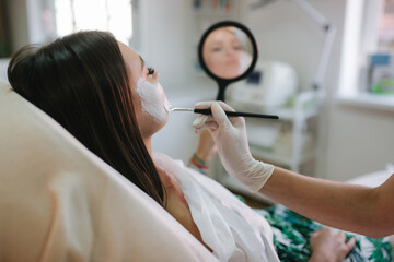 Hands of cosmetology specialist applying white facial mask using brush. Making skin hydrated and face glowing and skin. Hulf face with clay mask. Attractive young brunette relaxing with closed eyes