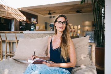 Pensive intelligent hipster girl in eyewear for vision correction holding book sitting on cozy interior, confident caucasian pretty woman writer signing autograph on her