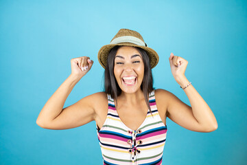 Young beautiful woman wearing swimsuit and hat over isolated blue background very happy and excited making winner gesture with raised arms, smiling and screaming for success.
