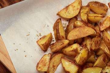 Part of white paper and cutting board with delicious baked golden potatoes slices with seasoning from top. On wooden table with free space for text