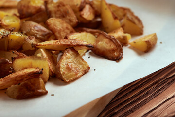 Part of white paper and cutting board with delicious baked golden potatoes slices with seasoning from sideways. On wooden table with free space for text