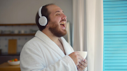 Happy man in bathrobe with cup of coffee listening to music in headphones standing near window