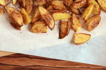 Part of white paper and cutting board with delicious baked golden potatoes slices with seasoning from top. On wooden table with free space for text