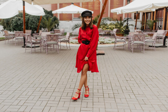 Girl in elegant, red belted dress and shoes on city heel shows slender legs. Full-length photo in city cafe