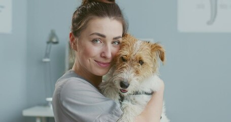 Close up portrait of female veterinarian holding dog and kissing it while waiting for owner after examination at clinic. Vet sitting in medical suit. Concept pets care, veterinary, healthy animals.