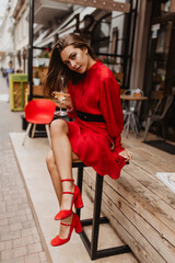 Photo in full growth of gorgeous brunette. Girl in scarlet dress romantic and cute smiles into lens while sitting in cafe. Woman enjoying good day while drinking wine