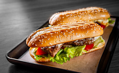 Two long submarine sandwiches with meat, cheese, bacon, tomatoes, lettuce, cucumbers and onions on black wooden table background