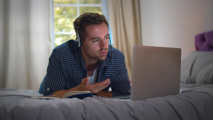 Young man working at home using laptop and headset lying on bed