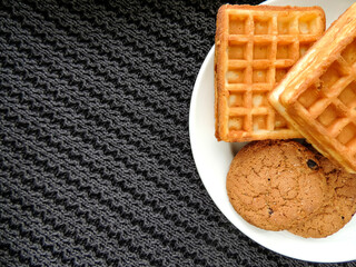 Homemade cakes on a black knitted background. Waffles and oatmeal cookies cooked at home on a white plate. Home-made food close-up and place for text.