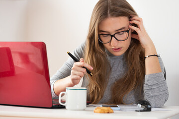 Natural blonde european woman doing her work remotely in front of red laptop at desk.  Isolation time. Pandemic threat. Virus outbreak implications. 